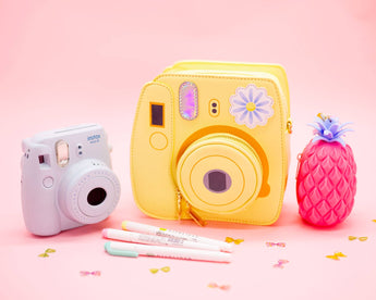 Mellow Yellow instant camera handbag displayed with a pink camera, pineapple and pens