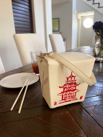 Cream Chinese takeout box crossbody bag displayed on a dining table with chopsticks